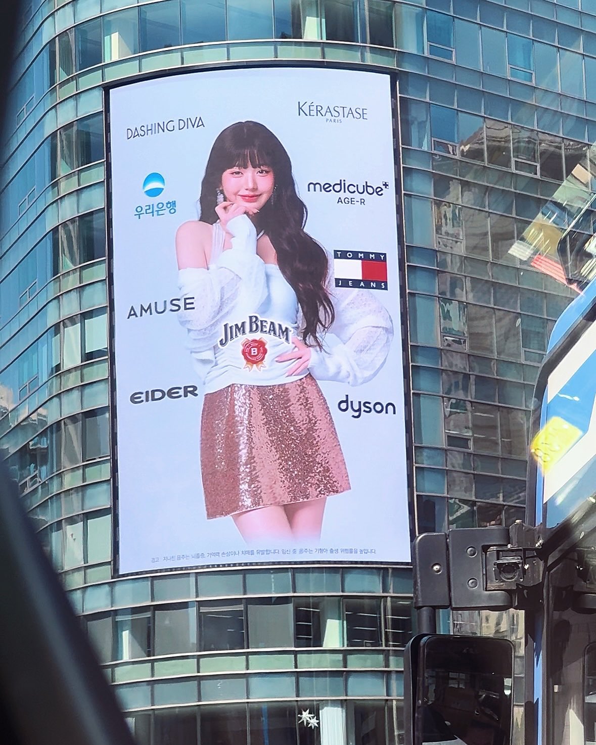Jang Wonyoung in a multi-brand commercial, elegantly posing with different brand products like a Dyson hair tool and a Jim Beam bottle, showcasing her versatility as a model for various industries.